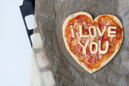 Italian heart shaped pizza with salami, tomato sauce, parmesan, pizza sauce, mozzarella and olive oil on parchment paper with cheese I love you. Love concept for Valentine's Day February 14