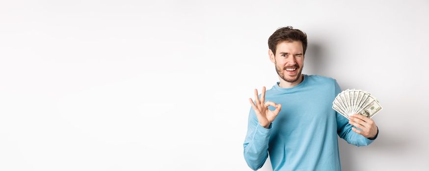 Cheeky smiling man winking, showing Ok sign and holding money, concept of fast loan or credit, standing over white background.