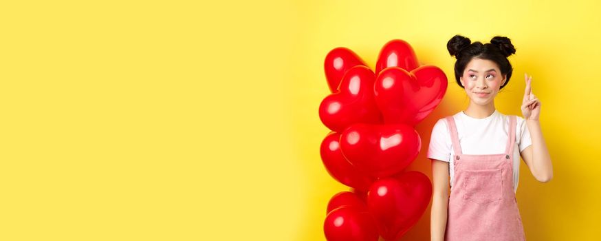 Valentines day concept. Beautiful korean girl dreaming of perfect date, making wish and looking at upper left corner, standing near red hearts balloons, yellow background.