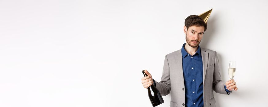 Party guy standing in birthday hat and celebrating, holding champagne bottle and glass, looking drunk, wearing grey suit.