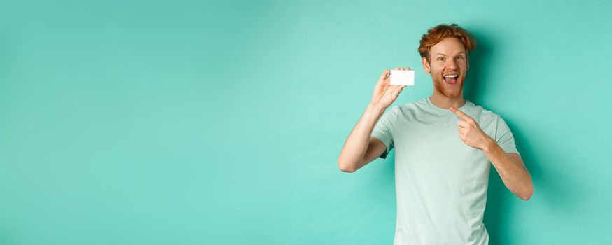 Shopping concept. Handsome redhead man in t-shirt showing plastic credit card and smiling, standing over turquoise background.