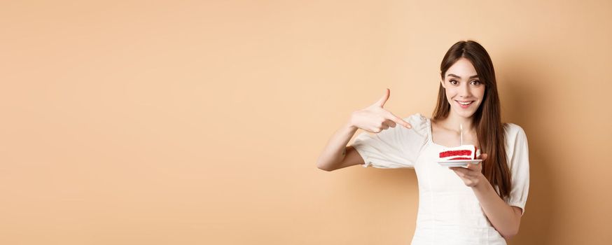 Cute girl pointing at birthday cake with candle and smiling excited, making bday wish, standing on beige background.