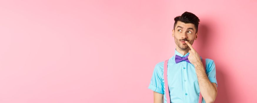 Image of handsome funny man in bow-tie looking thoughtful left and touching lip, thinking what to choose, making decision, standing over pink background.