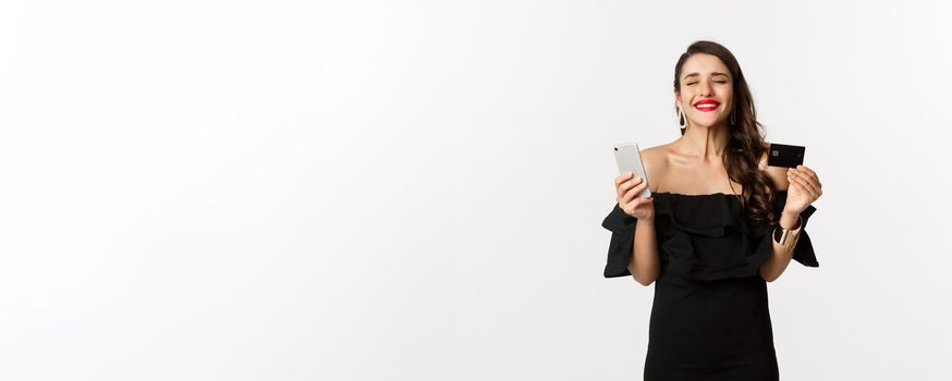 Online shopping concept. Fashionable woman in black dress, holding credit card with smartphone, looking satisfied, standing over white background.