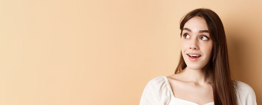 Dreamy beautiful girl looking at upper left corner logo, checking out banner, standing thoughtful on beige background.