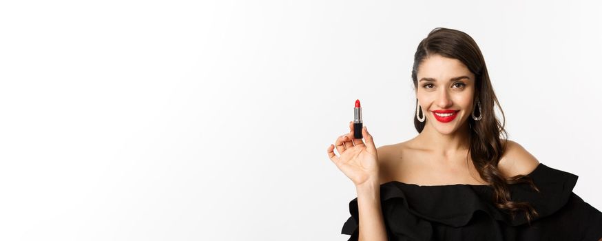Fashion and beauty concept. Beautiful woman in black dress showing red lipstick and smiling, standing over white background. Copy space