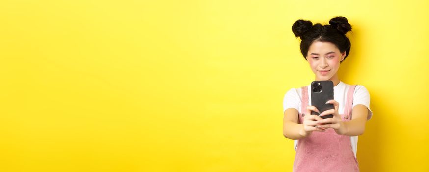 Stylish asian girl taking photo on smartphone, making video with cellphone and smiling, standing on yellow background.