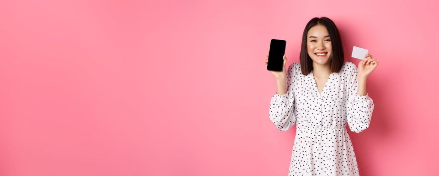Joyful korean woman showing smartphone screen and credit card, paying for internet order, demonstrating online shopping app, standing over pink background.