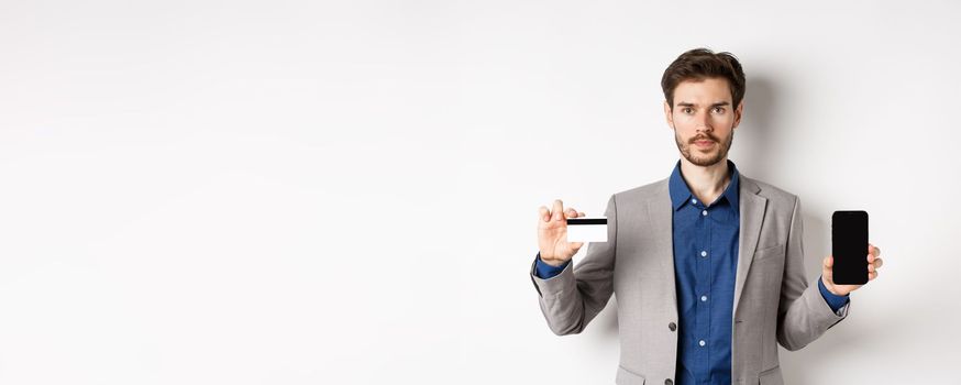 Online shopping. Serious bearded man in business suit showing plastic credit card with empty smartphone screen, standing against white background.