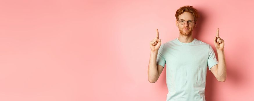 Confident and pleased young man with red hair, wearing glasses and t-shirt, pointing fingers up and smiling with smug face, showing good deal, standing over pink background.