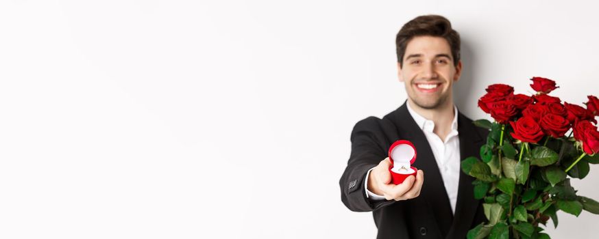 Close-up of attractive man in suit, holding bouquet of roses giving an engagement ring, proposing to girlfriend, standing against white background.