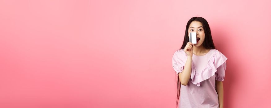 Excited asian girl showing plastic credit card and smiling, going on shopping, standing against pink background.