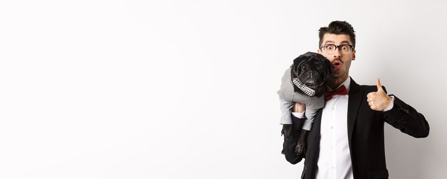 Happy young hipster in suit and glasses, showing thumb-up, holding cute black dog on shoulder, love his pug, standing over white background.