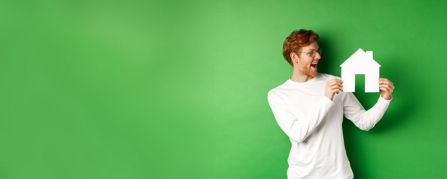 Real estate. Cheerful caucasian man with red hair, looking at paper house cutout and smiling amazed, buying property, standing over green background.