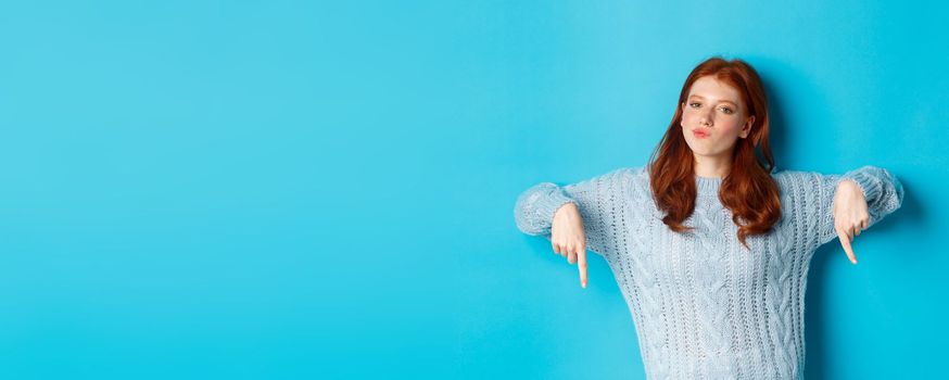 Winter holidays and people concept. Thoughtful teenage redhead girl in sweater, pointing fingers down and pondering, making decision, standing over blue background.