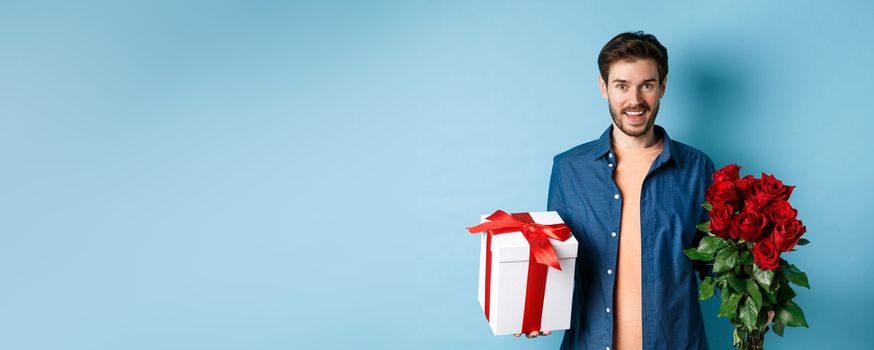 Love and relationship concept. Happy young man bring flowers and gift on romantic date. Boyfriend wish bouquet of roses and present standing on blue background.