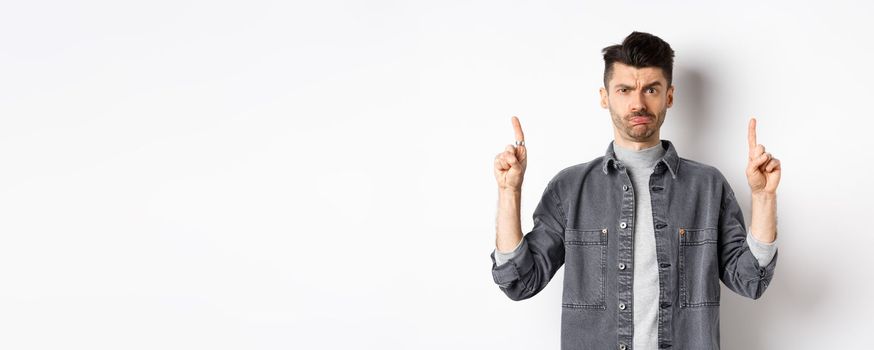 Confused funny guy with moustache pointing fingers up at something strange, frowning and pouting puzzled, standing on white background.