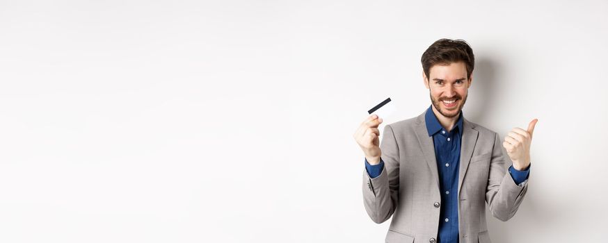 Happy bank client in business suit showing plastic credit card and thumb up, smiling satisfied, standing on white background.