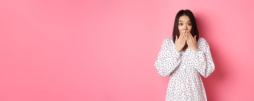 Worried asian girl in dress looking concerned, cover mouth and gasping, staring at camera shocked, standing in cute dress over pink background.