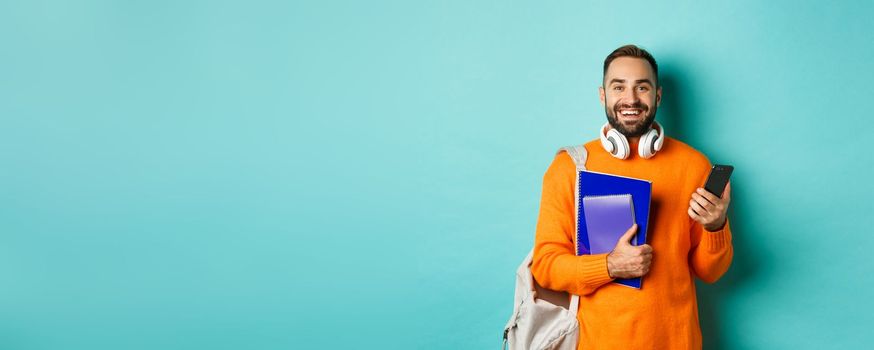 Education. Handsome male student with headphones and backpack, using smartphone and holding notebooks, smiling happy, standing over turquoise background.