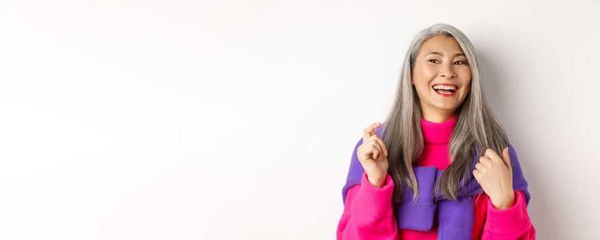 Close-up of fashionable asian senior woman, laughing and having fun, looking left cheerful, standing over white background.