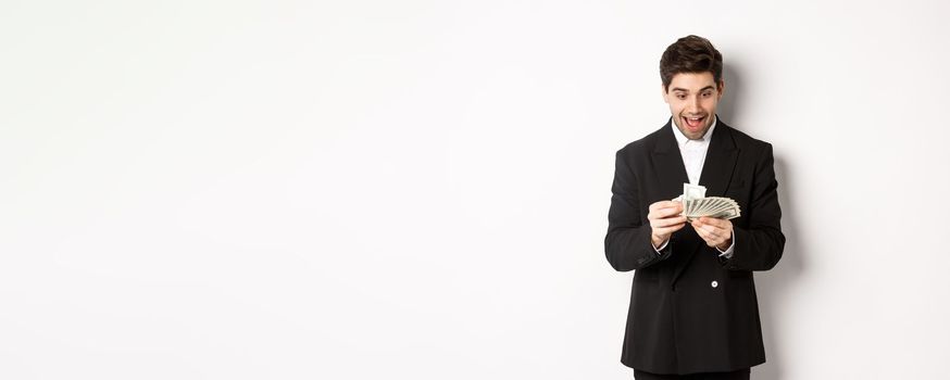 Image of excited handsome businessman, counting money and smiling amused, standing against white background in suit.