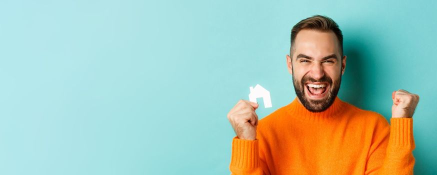 Real estate. Cheerful man buying apartment, rejoicing and saying yes, showing small paper house, standing over light blue background.