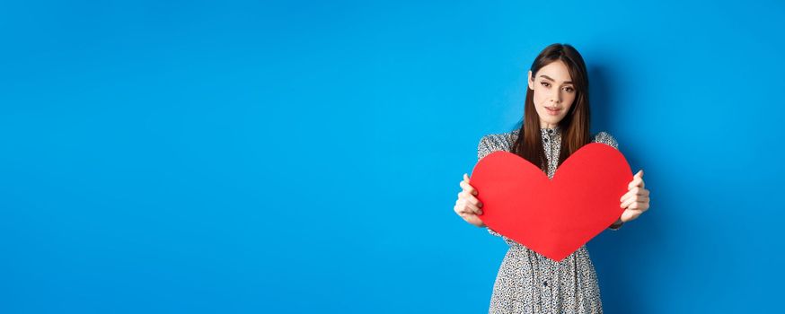 Valentines day and relationship concept. Tender young woman in dress stretch out hand and giving big red heart to you, making confession, standing romantic on blue background.