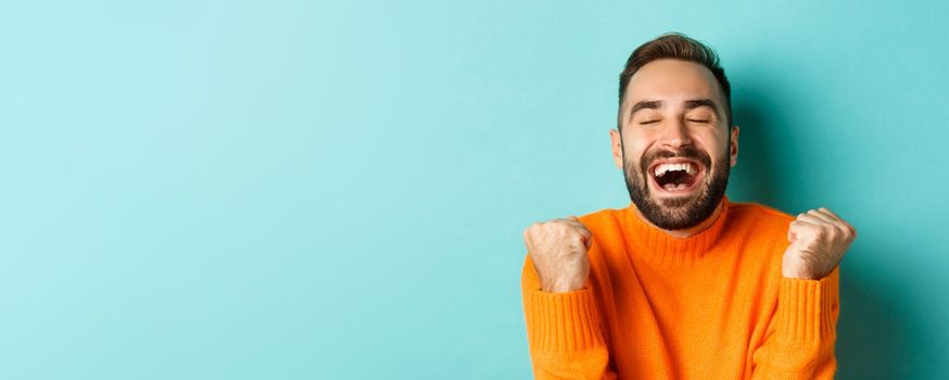 Image of handsome relieved man feeling satisfaction, rejoicing of winning or achievement, making fist pump and saying yes, standing over turquoise background.