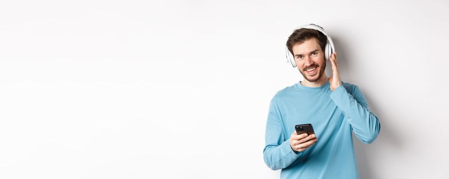 Good-looking guy listening music in wireless headphones, smiling at camera and using smartphone, standing on white background.