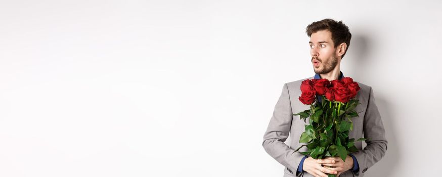 Nervous boyfriend waiting for his date on valentines day, looking left amazed, holding bouquet of red roses, standing in suit over white background.