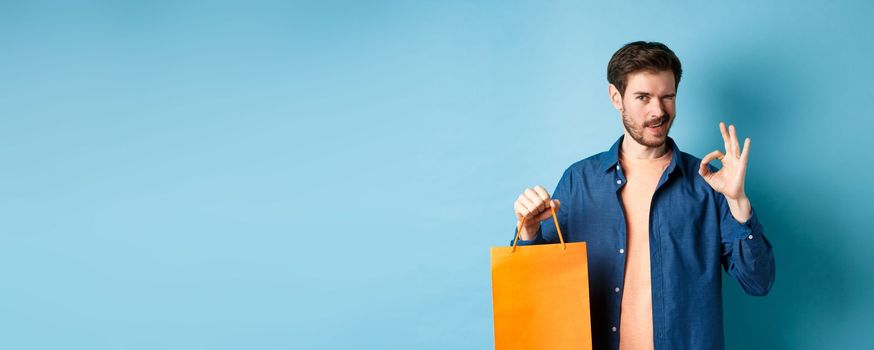 Modern man in casual outfit showing shopping bag and okay sign, winking at camera, recommending shop, standing on blue background.