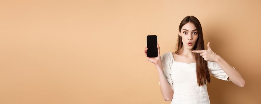 Excited woman showing news on screen, pointing at empty phone and looking surprised, standing on beige background.