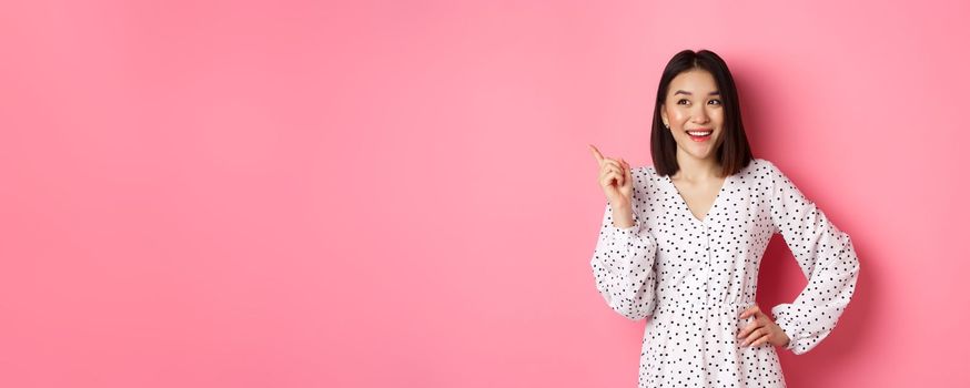 Happy asian girl in dress smiling satisfied, pointing finger at upper left corner, nod in approval, standing in dress against pink background.