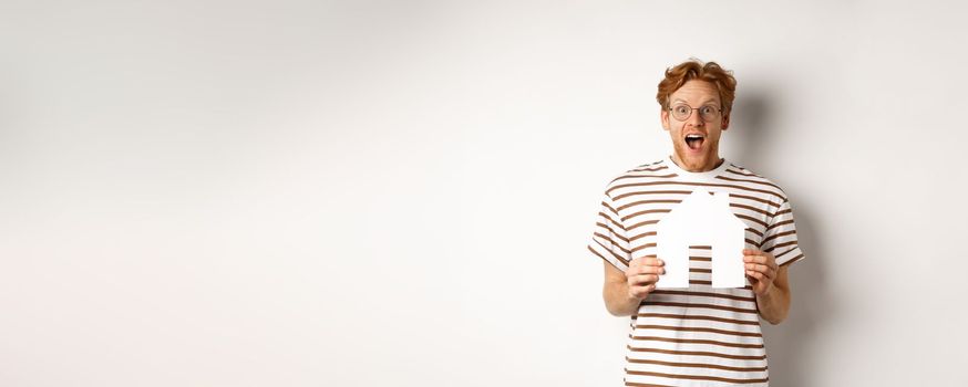 Happy and surprised redhead man winning house, holding paper home model and staring at camera, standing joyful over white background.