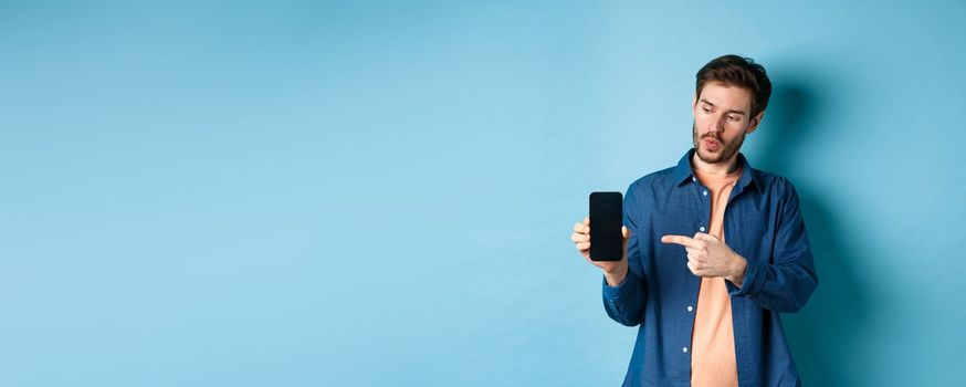 Image of handsome man talking about application, pointing finger at mobile screen and standing on blue background.
