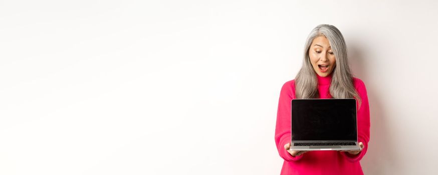 E-commerce concept. Amazed asian grandmother with grey hair, checking out promo online, showing laptop black screen, standing over white background.