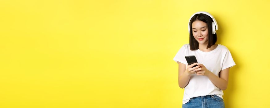 Modern asian girl listening music in wireless headphones, reading smartphone screen and smiling, standing in white t-shirt over yellow background.