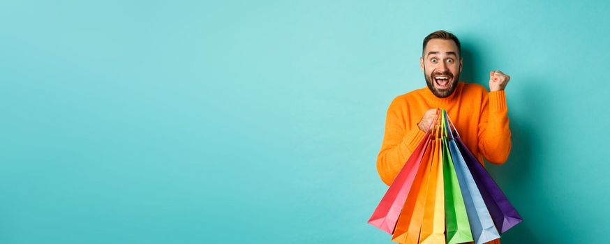 Lifestyle concept. Excited man showing shopping bags and rejoicing from discounts, standing over turquoise background.