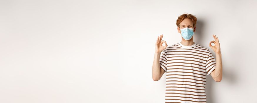 Covid-19, pandemic and social distancing concept. Young man with red hair wearing medical mask to prevent catching coronavirus, showing okay signs in approval, white background.