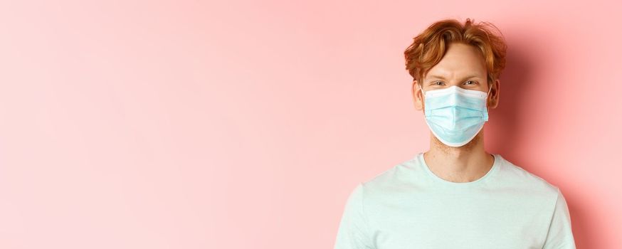 Covid-19 and pandemic concept. Close up of redhead man in medical mask looking at camera, standing over pink background.