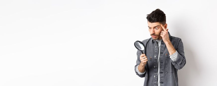 Man looking through magnifying glass and stretching eyelid to see clearly, standing on white background.