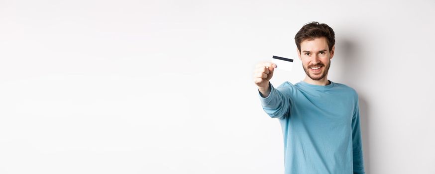 Confident young man smiling, stretch out hand and showing plastic credit card, standing on white background.