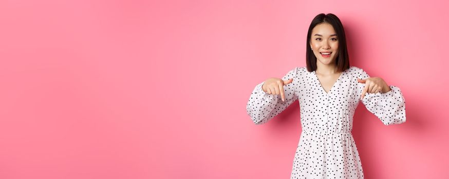 Look there. Cute asian woman in dress, pointing fingers down at copy space, showing product discount and smiling, standing over pink background.