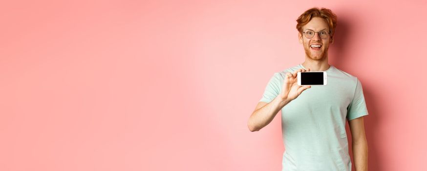 Happy young man with red hair showing smartphone screen, holding phone horizontally and smiling amazed, standing over pink background.