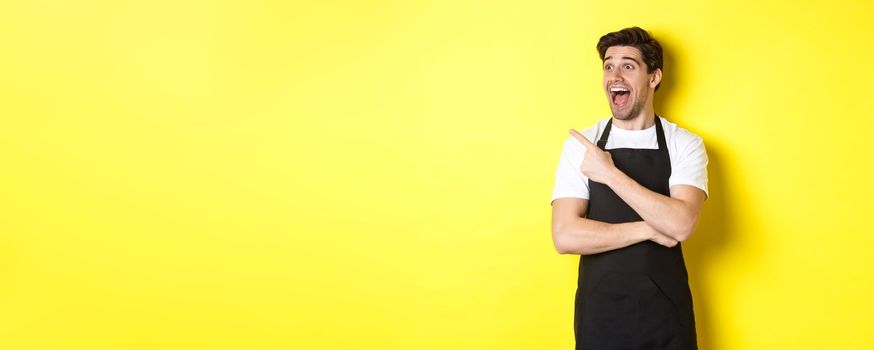Waiter in black apron checking out promo offer, pointing finger and looking left at your logo, standing over yellow background.