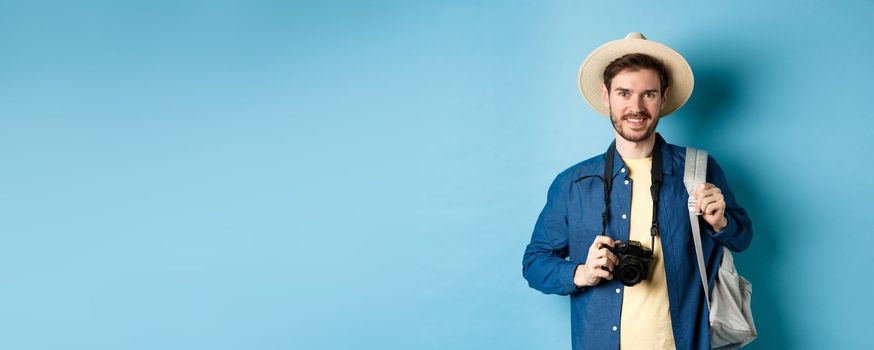 Cheerful handsome guy going on vacation, wearing summer hat and holding backpack with camera for photos, smiling excited of holiday, standing on blue background.