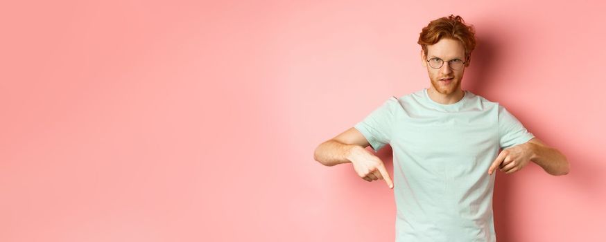 Confident redhead man in glasses pointing fingers down, staring daring with smug face at camera, showing advertisement, standing over pink background.