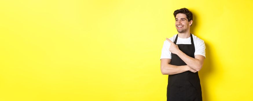 Handsome barista pointing and looking left at promo, wearing black apron, standing against yellow background.