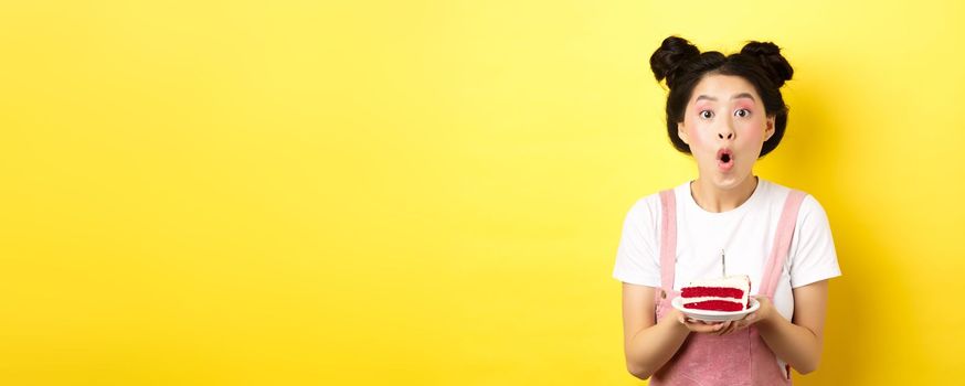 Happy asian birthday girl with bright makeup, blowing candle on cake, making wish, standing on yellow background.